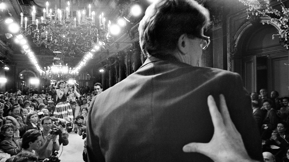 Imagen de la presentación de la colección de alta costura primavera-verano de Ives Saint Laurent en enero de 1986. El modista es obligado a salir al escenario para saludar a sus incondicionales. La mano que le empuja es la de su socio, amante y mentor Pierre Bergé, siempre entre bambalinas.